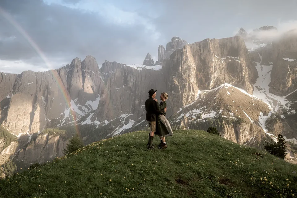 A couple in Tyrolean dress on the meadow at Passo Gardena, peaks and light behind
